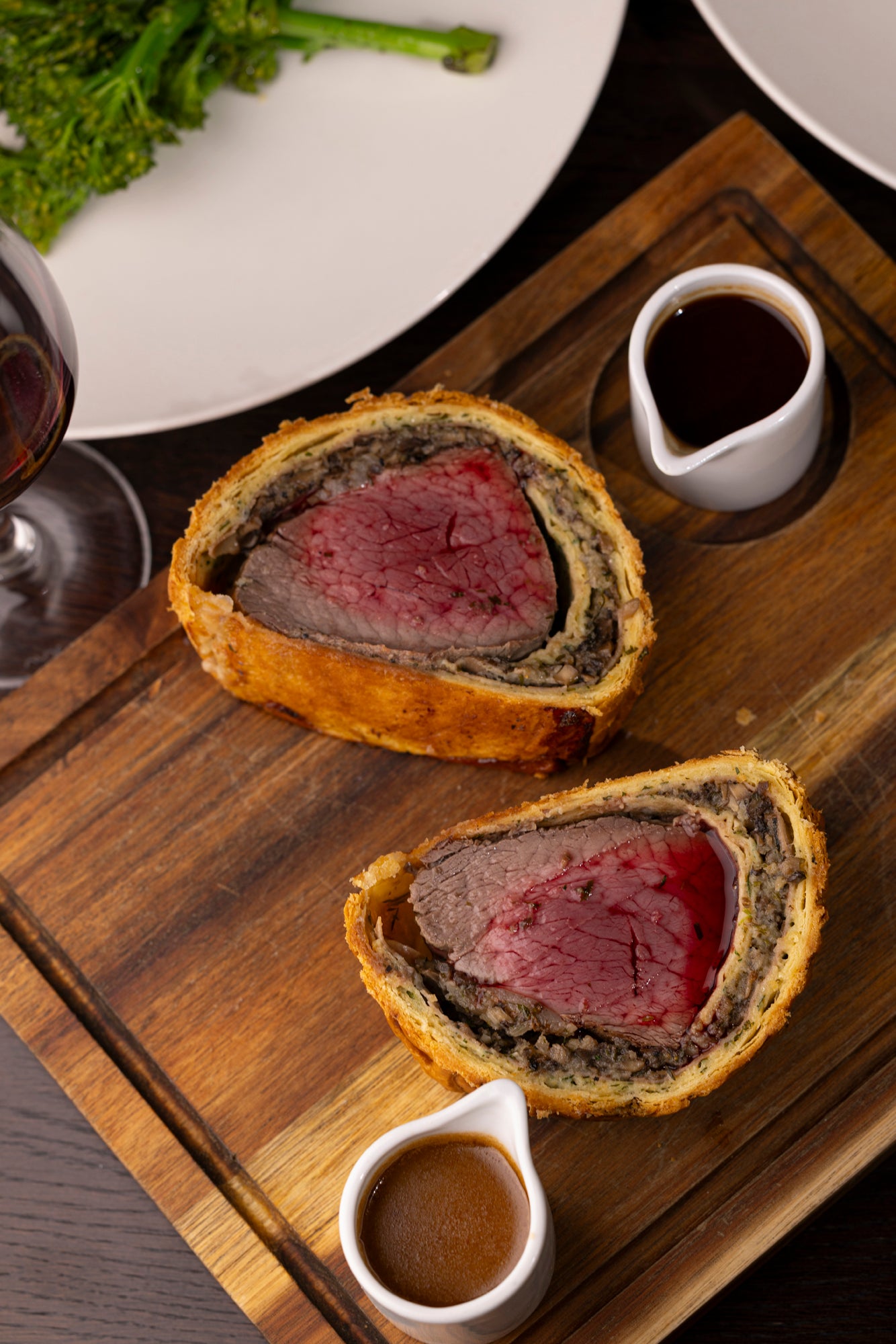 Two slices of beef Wellington on a wooden board with side dishes.